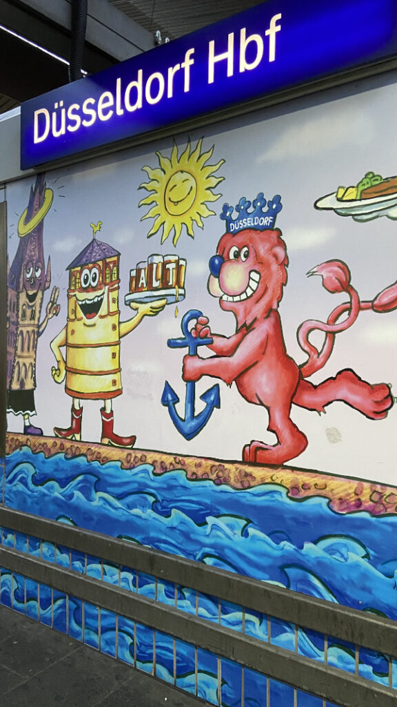 Eine Wandbemalung im Düsseldorfer Hauptbahnhof. Eine lächelnde Sonne, darunter ein Löwe, der Rheinturm mit Gesicht und einem Tablett Altbier in der Hand, daneben eine Kirche, ebenfalls mit Gesicht.