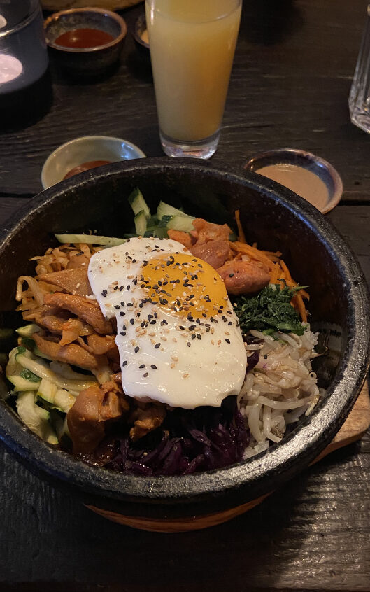 Ein koreanischer Hot Pot mit verschiedenem Gemüse und Fleisch, als Topping ein Spiegelei, serviert mit zwei Dips auf einem dunklen Holztisch.